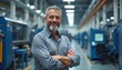 © miss irine - Man with grey beard smiles arms crossed in factory aisle. He oversees industrial production line with machines. This represents business owner, industry leader, manufacturing executive.