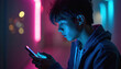 © miss irine - Young man looks at smartphone screen in dark room. Neon lights glow blue and pink. He is holding mobile device in his hands, focused and serious.