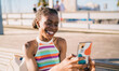 © BullRun - Happy African woman holding phone during video chat, laughing with genuine delight, capturing light-hearted digital connection and youthful self-confidence on the go.