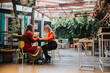 © qunica.com - Two women sit across from each other at a colorful outdoor cafe, talking over a table.
