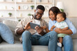 © Prostock-studio - Happy African American parents and their young son relax on a comfortable sofa, engaging with a tablet.
