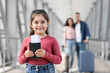 © Prostock-studio - A young girl smiles while holding her passport near the airport terminal. In the background, her parents are getting ready for their trip, showing excitement for travel.