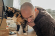 © kittyfly - bald man in brown sweater gently holding tortoiseshell cat on desk, working computer with keyboard and monitor, in cozy home office setting, petting cat during work break, furry companion nearby