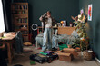 © DragonImages - Caucasian teenage girl standing in messy bedroom packing suitcase and cardboard box, organizing clothes and personal items, surrounded by books and plants, appearing thoughtful
