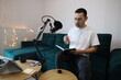 © YuliaSmyt - Asian man reviewing notes while recording audio at home, sitting by a microphone in a casual home studio. Focused remote-work or podcast preparation moment