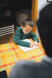 © Fotografia Juan Reig - Young boy in a green hoodie focused on writing in his workbook at a colorful kitchen table, learning and studying with a family member assisting him with school tasks