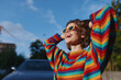 © SHOTPRIME STUDIO - Woman smiling in rainbow sweater and sunglasses, young adult laughing with hands in hair outdoors by parked car in sunny urban setting, joyful lifestyle fashion travel vibe