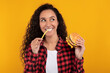 © Prostock-studio - A joyful woman bites into a delicious burger, holding a fry in her other hand. She looks excited, smiling against a vibrant yellow-orange background, ready to savor fast food.