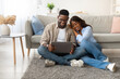 © Prostock-studio - In a warm and inviting living room, a young black couple sits on the floor, engaged with a laptop.