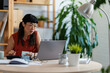 © lordn - Asian woman entrepreneur in her home office, checking receipts and entering numbers on her phone while working with a laptop and calculator at a round desk