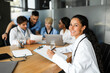 © Prostock-studio - Middle eastern woman doctor smiling at camera while having morning session with colleagues, multiracial group of physicians professional doctors discussing medical cases, using modern technologies