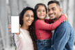 © Prostock-studio - A happy family stands together in a cheerful indoor environment. A woman smiles while holding a blank smartphone, while a man embraces a girl, who is also smiling joyfully.