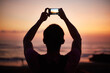 © pressmaster - Man holding smartphone above head capturing sunset over ocean, standing outdoors with arms raised, silhouetted against colorful sky during evening