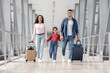 © Prostock-studio - A family walks hand in hand at the airport, carrying luggage. The adults and child appear excited as they head towards the departure gate.