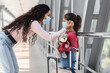© Prostock-studio - A woman helps her young daughter put on a mask at the airport. The girl holds a teddy bear, looking up at her mother. The airport interior is bright and modern.