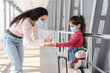 © Prostock-studio - A mother applies hand sanitizer on her daughter's hands while preparing for a journey at an airport. Both wear face masks and are surrounded by modern architecture.