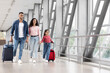 © Prostock-studio - A family of three is seen strolling through an airport terminal, holding hands and smiling. The parents are pushing wheeled luggage while their daughter plays with her smaller bag.