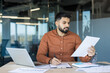 © Liubomir - Young male professional concentrating on official documentation while writing notes at a desk with a laptop and various business papers in a contemporary office setting