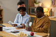 © pressmaster - Black young adult woman and Black senior man sitting at table reviewing financial documents and counting cash, using calculator and discussing paperwork related to paying taxes