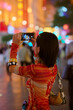 © Connect Images - Woman capturing colorful city lights at night on a smartphone, showing a bustling urban scene. Shanghai, China