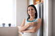 © Connect Images - Woman in a white top holding a glass, standing by a refrigerator with a confident smile. Germany