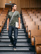 © Connect Images - Man in casual attire holding laptop, walking in an empty lecture hall. Germany