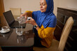 © Taras Grebinets - Contemporary Muslim Woman Working From Home Pouring Water Beside Laptop At Dining Table