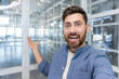 © Liubomir - Happy entrepreneur vlogging from a bright modern glass office, smiling and pointing as he introduces the open workspace to his online audience with enthusiasm and confidence