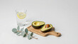 © LabioLoops - A clean, white countertop with a small wooden cutting board holding a halved avocado and a glass of clear water with a lemon slice. A single stem of eucalyptus lies beside it.