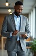 © Viktor - Black man uses a tablet at work. Confident ceo in suit stands in office. Manager works on digital device. Businessman checks email using portable gadget for online business.