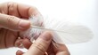 © RisaFitri - Close-up of hands holding a delicate white feather against a bright background.