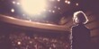 © Yuliia - Silhouette of female speaker facing audience in theater auditorium with dramatic stage lighting and blurred crowd in background.
