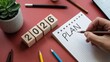 © MediaStockAI - Wooden blocks displaying the year 2026 rest beside a spiral notebook where the word plan is being written by a hand