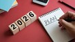 © MediaStockAI - Wooden blocks displaying the year 2026 sit beside a notepad where the word plan is being written