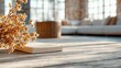 © shaki - A close-up of a closed book and dried floral arrangement on a weathered wooden table. In the blurred background, a large sectional sofa and a wicker basket are