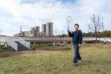 A young man is holding a badminton racket and standing on a grassy area in an outdoor park with buildings in the background.