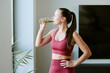 © AnnaStills - Young adult Caucasian woman standing near window drinking green smoothie with eyes closed, wearing sportswear, hand on hip, enjoying healthy morning routine after workout