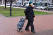 © Anna - Elderly woman on street. Pensioner walks with cart for things. Woman walks on road. Transportation of personal belongings.
