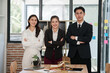 © aekachai - A group of three professional lawyers or legal consultants standing with arms crossed and smiling confidently in a modern office. On the wooden table in front of them are a scales of justice, a gavel,
