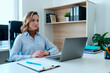 © Mediaphotos - Caucasian young adult woman sitting at desk using laptop, hands folded, looking slightly to side, working in modern office environment, documents and pen on table, bookshelf in background