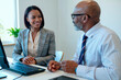 © Mediaphotos - Caucasian middle aged man and Black young adult woman sitting at desk discussing business matters in modern office setting, both engaged in professional conversation near desktop computer