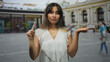© Krakenimages.com - Young latina woman points index finger with one hand and holds other palm up in front of an ornate building on a busy street; doubt choice decision.