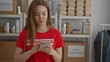 © Krakenimages.com - Woman wearing red volunteer shirt taps smartphone before stacked donation boxes in a building; charity compassion.