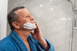 © svetlana_cherruty - Man in blue robe applies shaving cream in bathroom. Morning Routine. Side view. Close up.