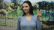 © Krakenimages.com - Young hispanic woman smiling and making a peace sign in an outdoor park setting, surrounded by lush greenery and a serene ambiance, conveying a sense of happiness and tranquility.