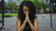 © Krakenimages.com - Young woman praying outdoors on a city street with blurred green background, eyes closed, hands together, expressing peace and tranquility, wearing black sleeveless top and glasses.