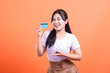© Ajab - A smiling young woman holds up a credit card, pointing towards it with a pleased expression, ready to spend her money, isolated on orange background