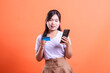 © Ajab - A young woman holding a credit card and using her smartphone for online banking, showing a modern and convenient way to manage finances, isolated on orange background.