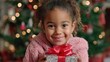 © mila103 - A joyful young girl stands holding a colorful present wrapped in shiny paper. Behind her a beautifully decorated Christmas tree is adorned with lights and ornaments creating a festive atmosphere.