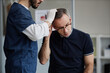 © Seventyfour - Middle aged Caucasian man receiving scalp examination from male healthcare professional wearing gloves during dermatology appointment, patient tilting head forward for inspection